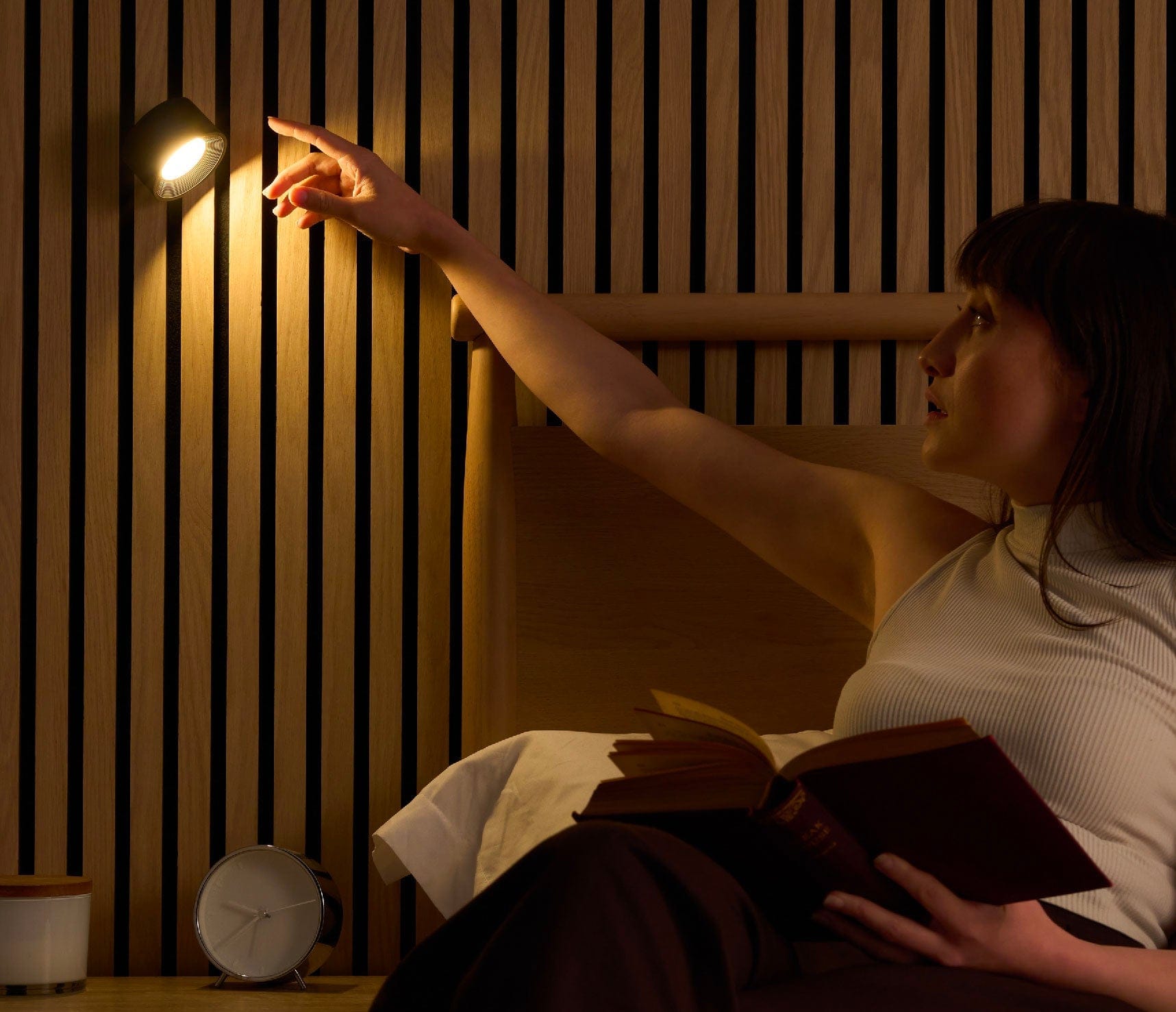 Person reading a book in bed with a night light on a wooden slatted wall.